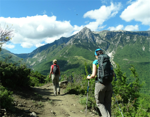 Two hikers with backpacks trekking a rocky trail through lush green valleys in Albania’s Accursed Mountains