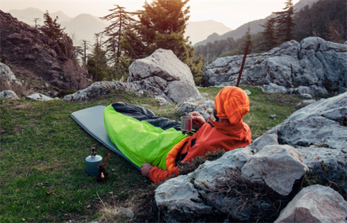 Hiker relaxing in a green ultralight bivvy bag with a cup of tea at sunset, demonstrating lightweight backcountry emergency shelter