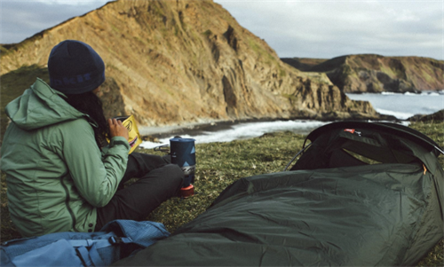 Solo camper with a green bivvy bag tent on a coastal cliffside, preparing a meal while using ultralight emergency shelter gear