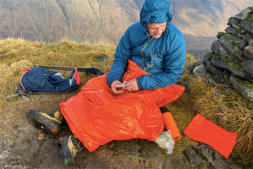 Mountaineer using an orange emergency bivvy bag (space blanket) on a mountain summit, showcasing survival gear in remote conditions