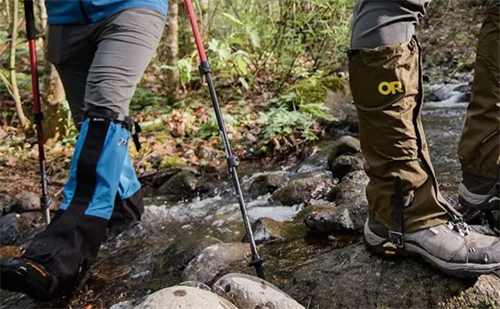 Waterproof hiking gaiters (including Outdoor Research models) protecting boots while crossing a forest stream on rocky trails