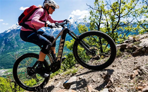 Female rider on a Trek electric mountain bike descending a rocky alpine trail, with snow-capped mountains in the background