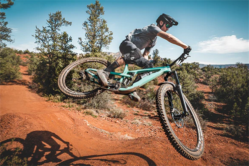 Rider performing a jump on a teal electric mountain bike on red desert singletrack, illustrating e-bike capabilities on trails