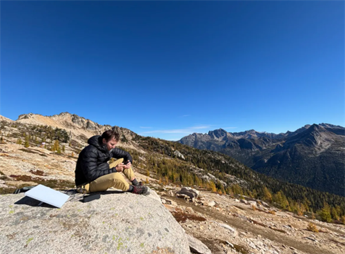 Hiker using a smartphone on a mountain summit with a Starlink dish nearby, showing high-speed internet access in remote alpine terrain