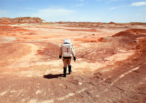 Hiker in a spacesuit-style outfit trekking across a red, Mars-like desert landscape, inspired by space tourism and off-world exploration