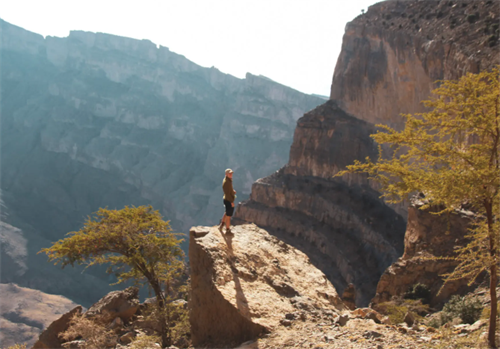 Trekking in Oman’s Hajar Mountains, a hiker stands on a rock outcrop with sweeping canyon views