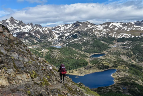 Solo hiker trekking a rocky slope with views of glacial lakes and snow-capped peaks on the Dientes de Navarino Circuit, Chile