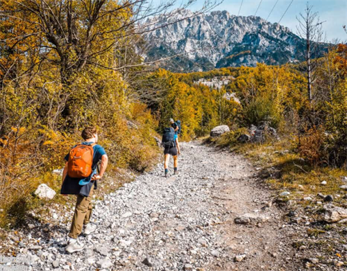 Two hikers trekking an autumn trail toward the snow-capped Albanian Alps on the Valbona–Theth route