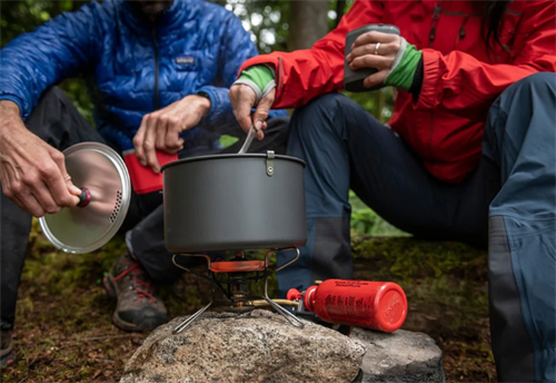 Two trekkers cooking with a liquid fuel stove system at a forest campsite, comparing different backcountry cooking setups