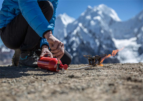 Hiker lighting a liquid fuel stove with a priming flame at high altitude, with snow-capped mountains in the background